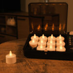 12 vandtætte genopladelige LED-fyrfadslys i sort opladningskuffert på træbord – ét lys udenfor, med stemningsfuldt rum i baggrunden