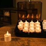 Et sæt med tolv flammeløse LED-lys og en fjernbetjening står i en åben sort kasse på et træbord med et lys placeret uden for kassen. Et hyggeligt, blødt oplyst rum er i baggrunden.