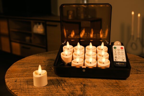 Et sæt med tolv flammeløse LED-lys og en fjernbetjening står i en åben sort kasse på et træbord med et lys placeret uden for kassen. Et hyggeligt, blødt oplyst rum er i baggrunden.