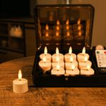 På et rundt træbord står et sæt flammeløse LED fyrfadslys og en fjernbetjening. Elleve lys er i et sort etui, og et lys står på bordet foran etuiet.