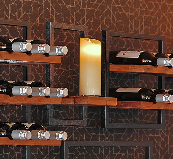 Ein modernes Weinregal mit Holzregalen und schwarzen Rahmen fasst mehrere Weinflaschen. Auf einem Regal steht eine brennende Kerze ohne Flamme. Der Hintergrund besteht aus einer geometrischen, braun gemusterten Tapete.