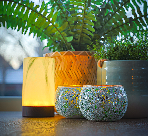 Eine leuchtend gelbe Lampe steht auf einem Tisch neben zwei Kerzenhaltern aus Mosaikglas. Im Hintergrund stehen ein geflochtener Korb mit einem grünen Farn und ein Keramiktopf mit einer anderen Pflanze.