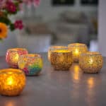 Twaalf waterdichte, oplaadbare waxinelichtjes met afstandsbediening (brandduur 175 uur, warm wit) gloeien in mozaïekglazen houders met gouden en kleurrijke patronen. Op de onscherpe achtergrond staan bloemen en een gezellige woonkamer.