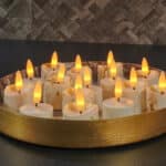 En rund, gylden bakke indeholder flere LED-fyrfadslys med 3D-flamme, brændetid 175 timer, genopladelige, med fjernbetjening, 6 stk., timerfunktion, arrangeret på en mørk bordplade med en flisebagvæg med struktur i baggrunden.