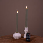 To tændte, olivengrønne LED-dinerlys (sæt med 2) står i hvide keramiske holdere på et træunderlag. Foran ligger en lille sort Dorsetto-fjernbetjening, med en ensfarvet brun baggrund, der skaber en rolig og varm stemning.