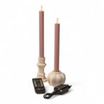 To lyserøde LED middagslys i hvide holdere med fjernbetjening og sort strømadapter på hvid baggrund, med realistisk flammeform.