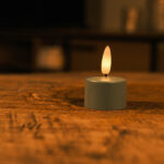 Sæt med 12 olivengrønne LED-fyrfadslys med fjernbetjening står på en træoverflade og giver et varmt, blødt skær i op til 400 timer, hvilket skaber en hyggelig og intim stemning.